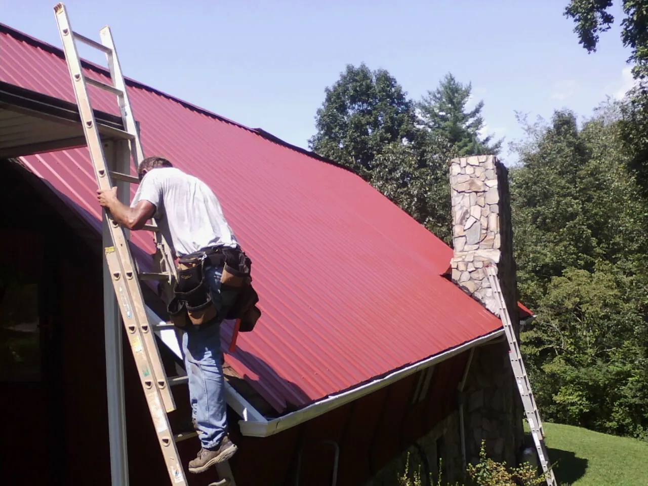 Roofer on a ladder beside a red metal roof and stone chimney — Soffit & Fascia Repair by Roofing Brothers in Port Norris